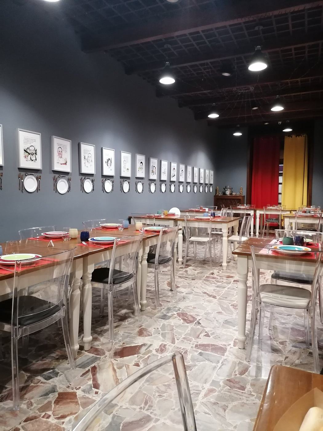 La sala di El Prado: parete grigia con caricature di chef incorniciate e piatti appesi, tavoli apparecchiati con piatti colorati, tende rosse e gialle in fondo